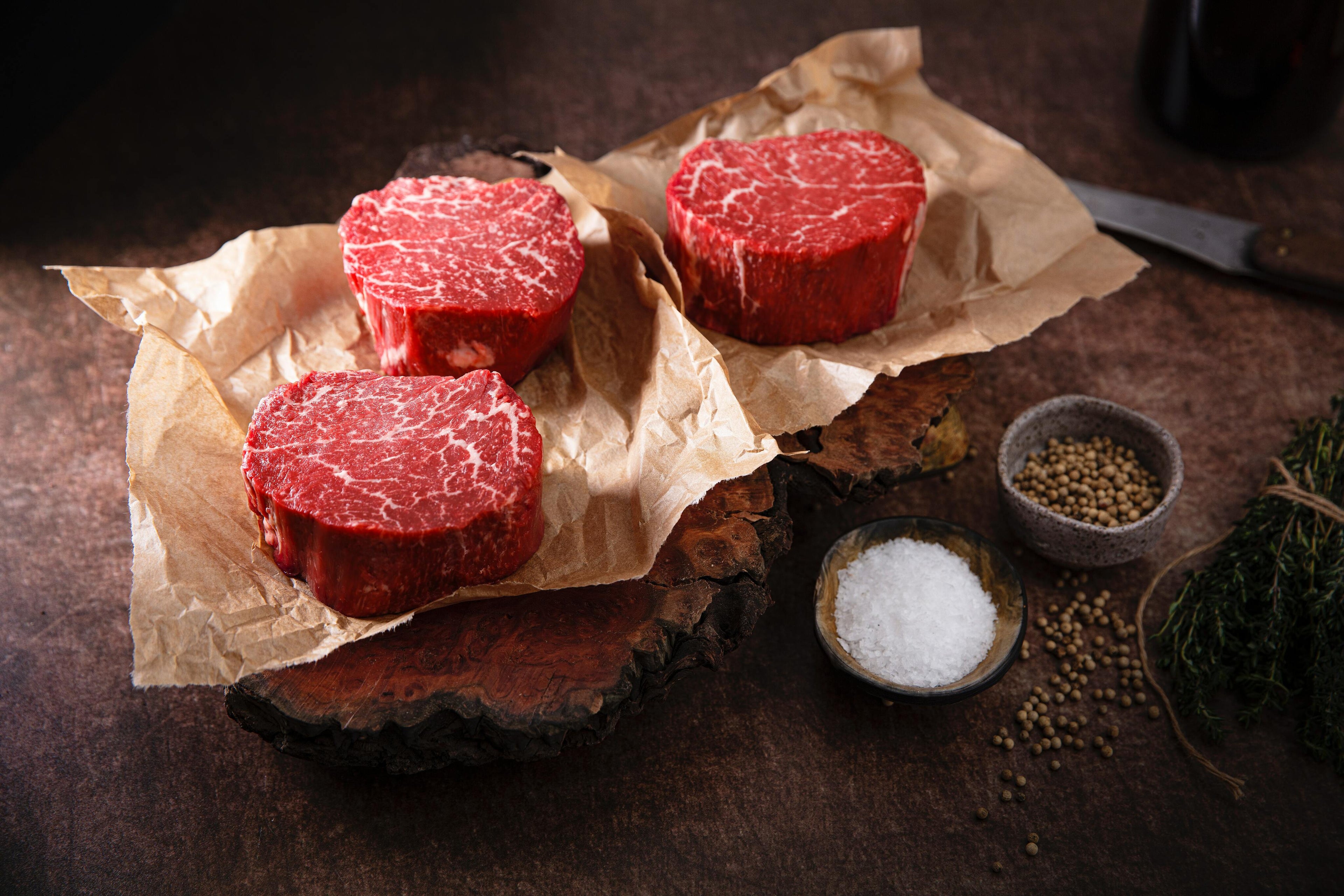 Several raw steaks on a piece of butcher paper with seasonings and a steak knife beside them, set on a wooden surface.